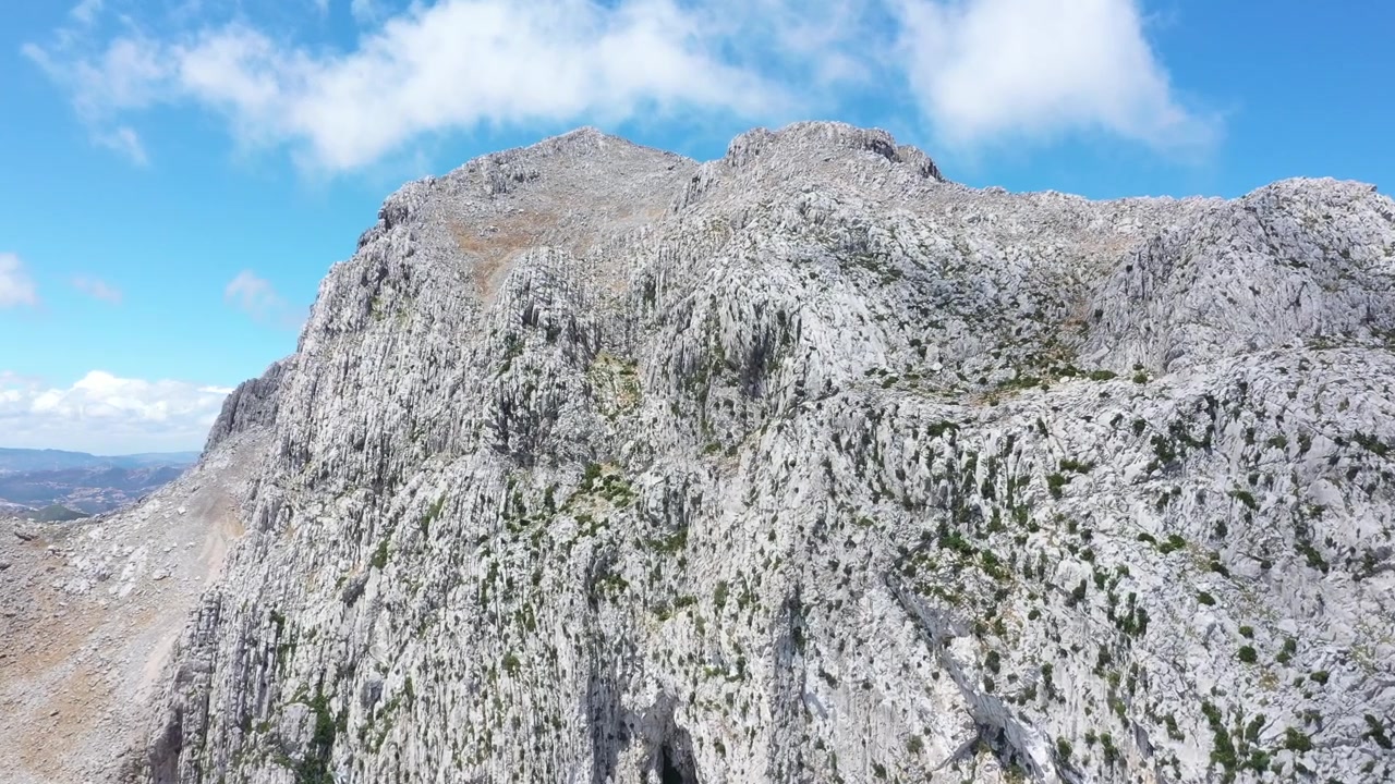 Vista aérea cercana de una enorme y escarpada formación montañosa en el norte de Marruecos, mostrando paredes rocosas expuestas, pendientes pronunciadas, vegetación escasa y texturas geológicas dramáticas bajo un cielo azul.