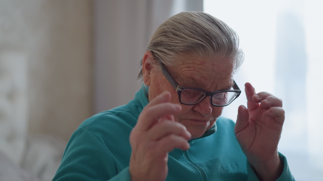 Elderly woman in teal green sweater sitting on bed, adjusting glasses with a thoughtful expression, soft natural light streaming through window, calm and peaceful home setting with minimalistic decor