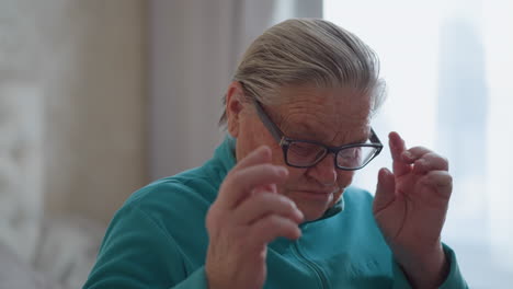 elderly woman in teal green sweater sitting on bed, adjusting glasses with a thoughtful expression, soft natural light streaming through window, calm and peaceful home setting with minimalistic decor