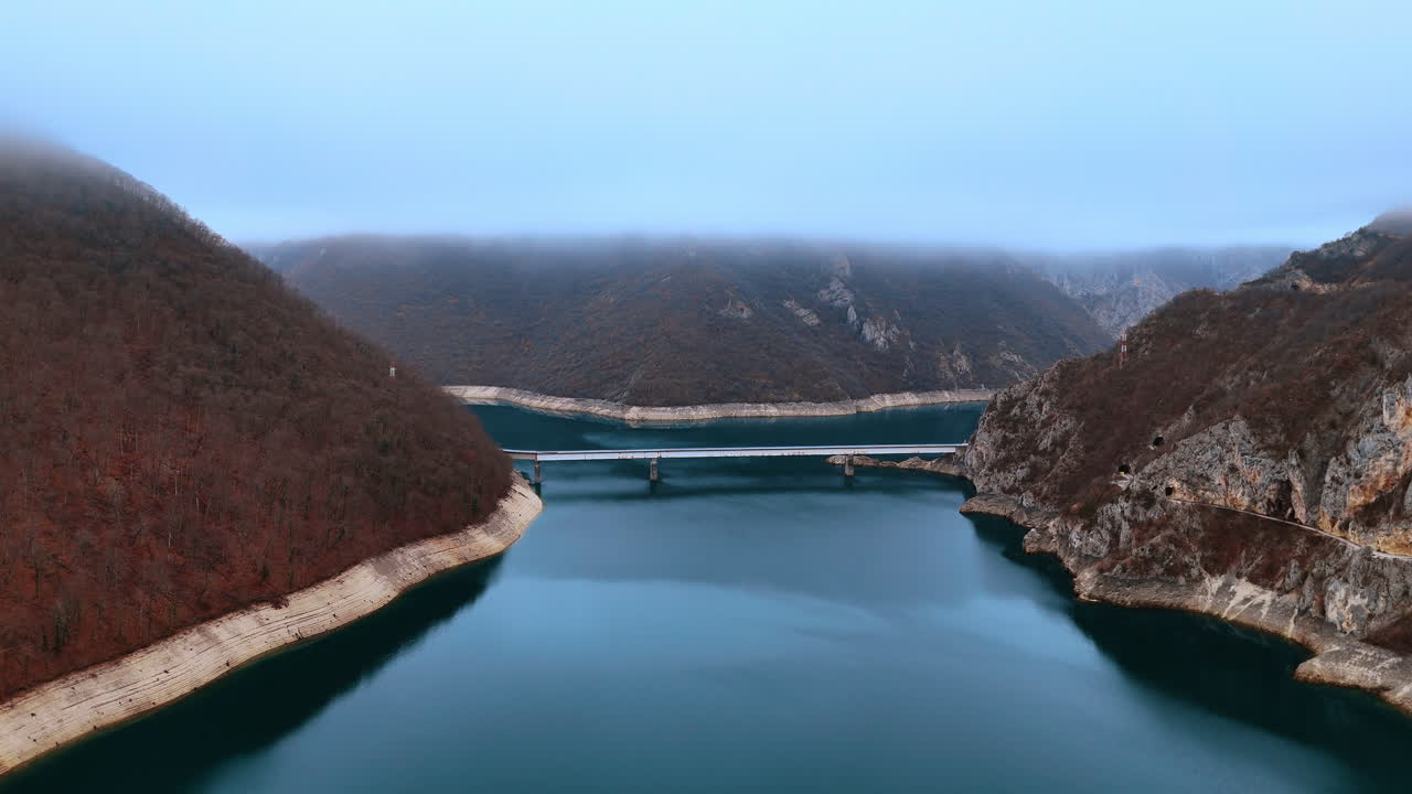 Wide aerial shot of bridge over Piva Lake canyon. Panoramic drone view of mountain bridge crossing turquoise reservoir under thick low clouds and mist