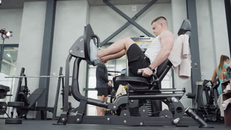 gym enthusiast using leg press machine while instructor exercises in background under bright gym lighting showing focused lower body strength, intense controlled motion and proper form