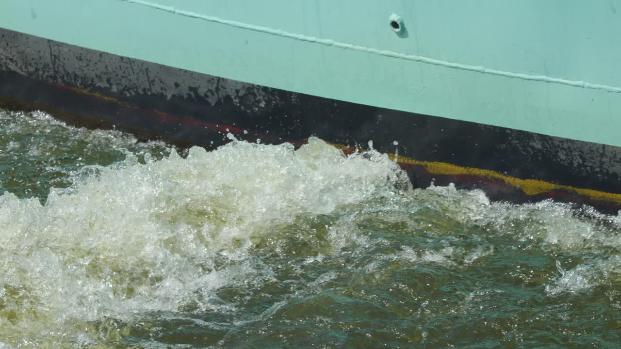 Close-up view of a boat hull as its engine agitates river water, producing foamy waves and turbulence under natural daylight with a static camera angle