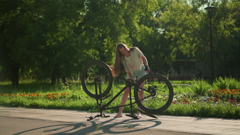 young woman rotates front tire and pedal of upside-down bike near vibrant garden, sunlight illuminates green park setting as blurred figure walks in background