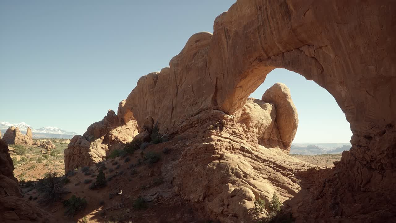 Pan right handheld wide landscape shot of large stunning natural orange sandstone arch formations with sun shining through them on a hike in southern Utah on a sunny warm spring morning
