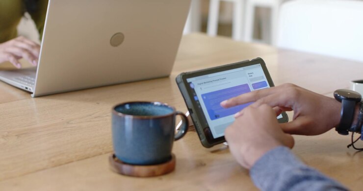 As assistant is propping tablet, tablet hands are tapping and moving content near laptop, reviewing