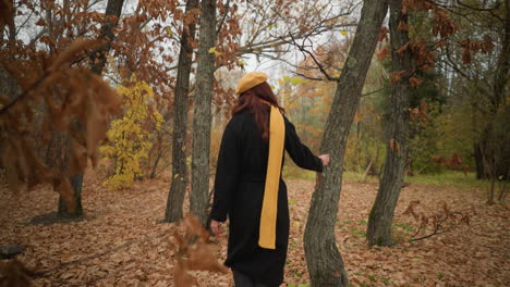 back view of a woman in a black coat and yellow muffler walking through a forest, touching dry leaves and tree bark, immersed in nature