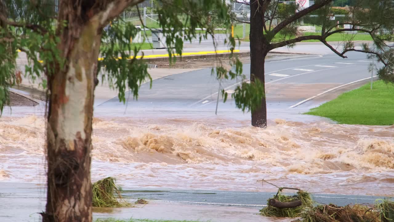 Wide shot of a flooded river on the Gold Coast, Queensland, Australia. Brown water flows rapidly over its banks, surrounded by green grass and trees under cloudy skies.