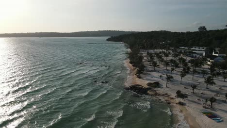 Kurze-Wellen-Rollen-Auf-Dem-Weißen-Sand-Mit-Kleinen-Palmen-Auf-Koh-Rong-Sanloem,-Während-Die-Sonne-Gerade-Aufgegangen-Ist-Und-Das-Licht-Auf-Dem-Meer-Reflektiert-Wird