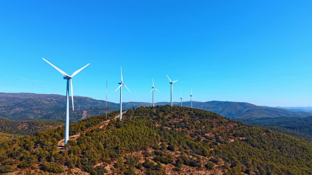 Alicante, Spagna, 8 agosto 2025: Turbine eoliche su colline boscose per la produzione di energia rinnovabile. Fila di moderni generatori eolici su una cresta montuosa sotto un cielo azzurro limpido in Spagna.