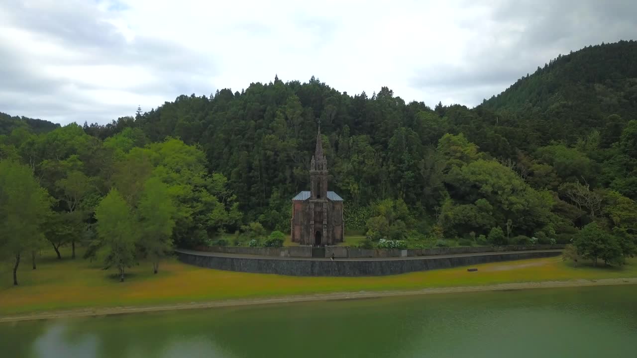 Drone flying backwards revealing old church made of stone in lake banks in Sao Miguel island, Azores.