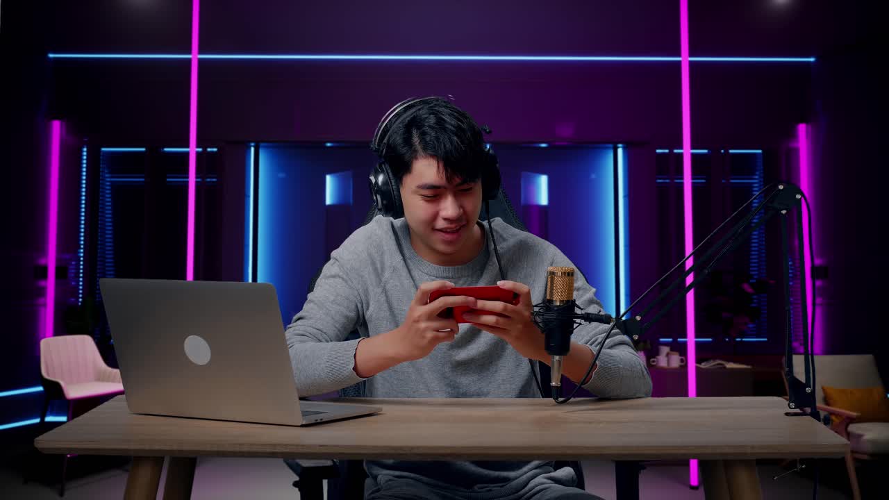 un jugador asiático sonriente con auriculares y computadora mirando a la cámara mientras usa el teléfono móvil jugando al juego en una habitación iluminada por luces de neón en retro