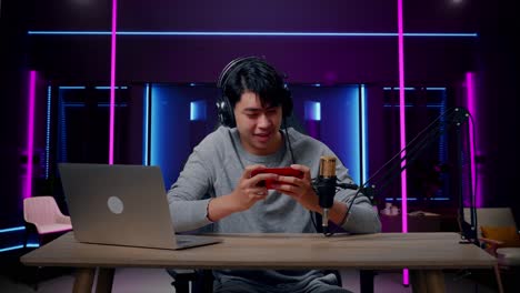 um jogador de videogame asiático sorridente com fones de ouvido e computador olhando para a câmera enquanto usa telefone celular jogando jogo na sala iluminada por luzes de néon em retro