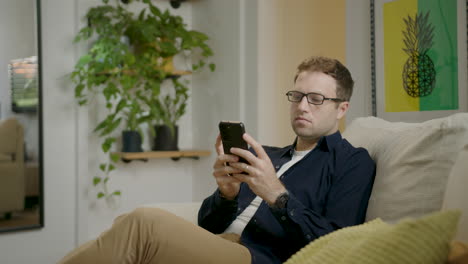 Serious-Caucasian-Man-Sitting-On-Couch-And-Texting-On-Smartphone-At-Home