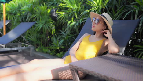 a young woman in a one-piece yellow bathing suit, sun hat, and sunglasses relaxes on a sun lounger around the pool