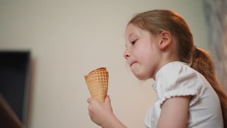 little girl in white blouse eating ice cream cone indoors, enjoying her treat with a soft smile, side view shows television on wall and relaxed home setting