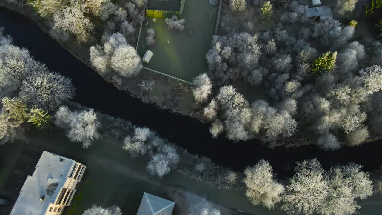 Aerial drone top down view flying over a small dark and cold urban small city river with some houses and roads around it with frost and hoard snow covered trees during morning time. Fresh white snow