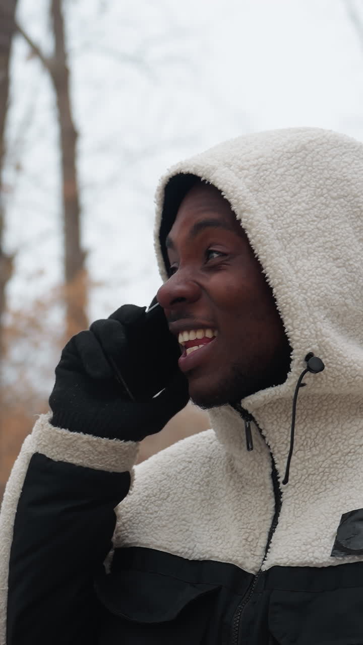 joven conversando por teléfono, sonriendo y participando en una conversación animada, con una sudadera blanca y guantes negros, con un fondo borroso de árboles, postes de luz de la calle en un entorno urbano