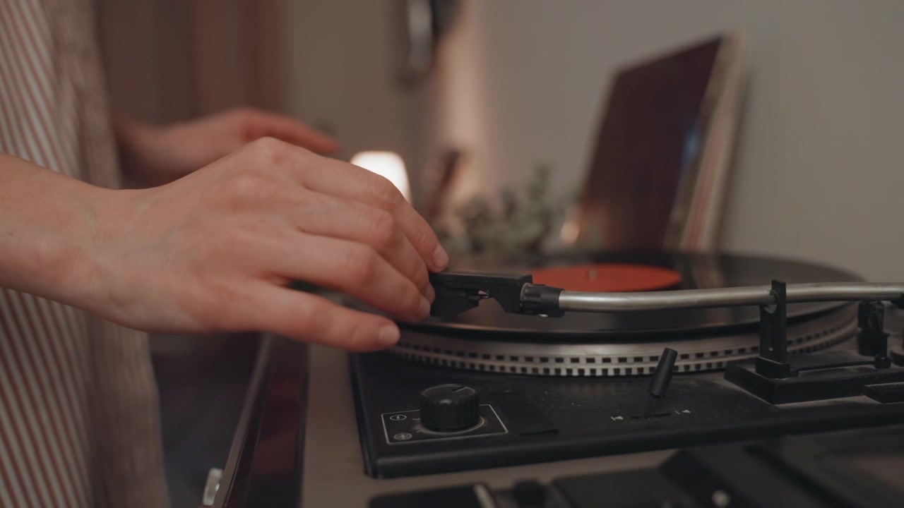 Donna caucasica amante della musica che tiene un vinile, posiziona il disco sul giradischi con le cuffie al collo, luce della lampada di un accogliente soggiorno, primo piano delle mani che regolano la puntina e il braccio, calore analogico vintage.