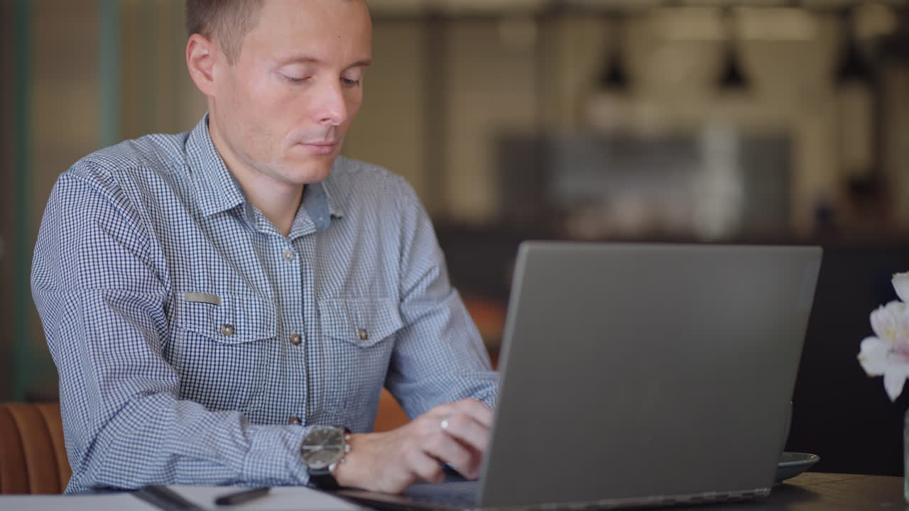 A young man in a shirt is sitting at a table with a laptop and typing on the keyboard. A student can study remotely. A businessman conducts his business remotely