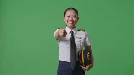 mujer piloto asiática sonriendo y tocando su pecho luego señalándote mientras está de pie en el estudio de fondo de la pantalla verde