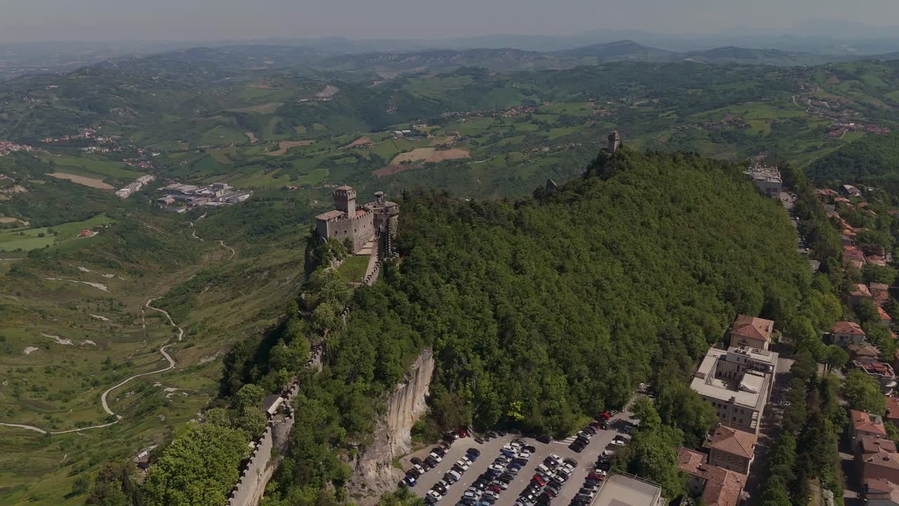 Drone shot of San Marino’s Cesta Tower (Seconda Torre) on Mount Titano. Dense green forest, dramatic cliffs and wide landscape views across the surrounding hills and countryside.