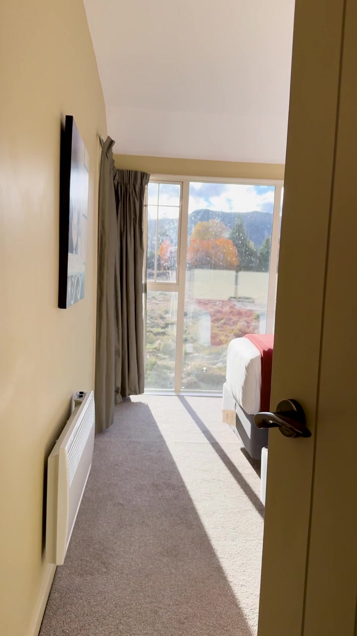 Una telecamera apre dolcemente una porta della camera da letto, rivelando una luminosa camera d'albergo illuminata dal sole con ampie finestre che si affacciano sul Lago Tekapo e su un paesaggio montano.