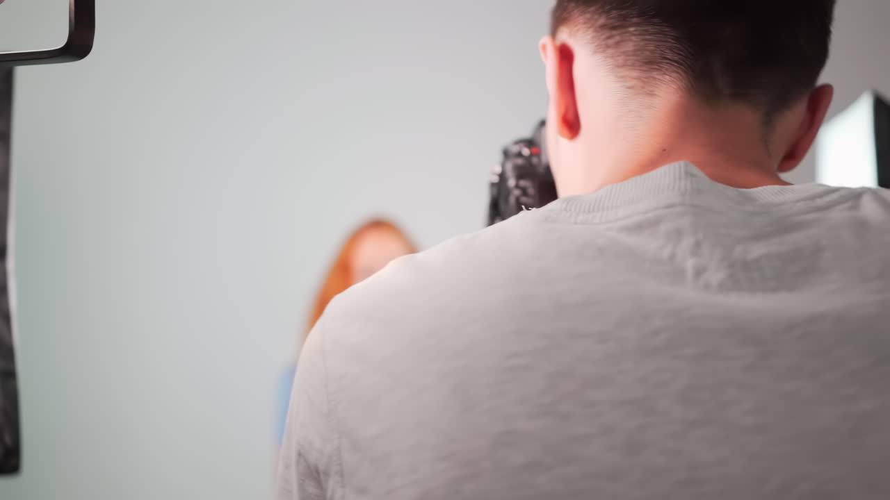Young professional photographer taking photos of a red haired girl in a studio with white background. Rear view. Girl with lots of freckles on the face