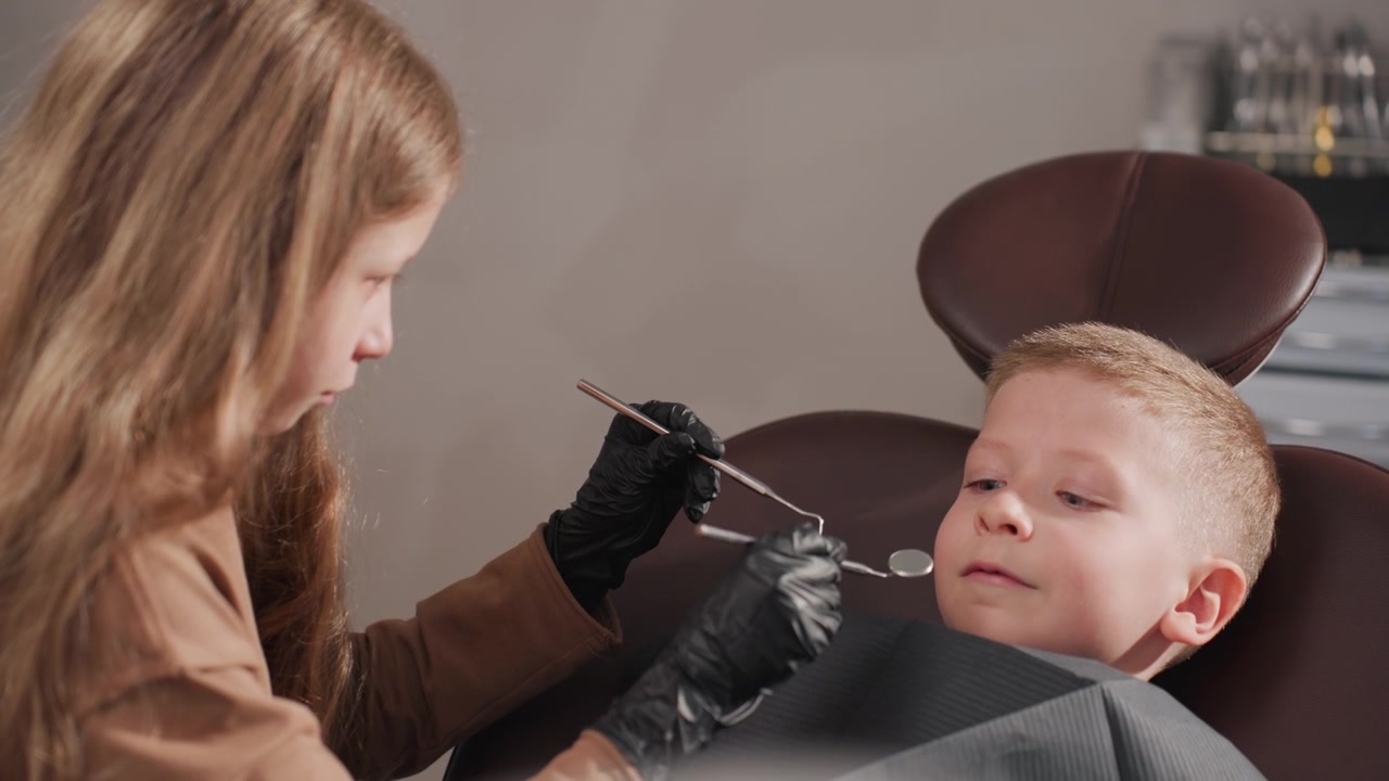 Primo piano di un bambino sdraiato su una poltrona da dentista mentre una dentista esamina i suoi denti con strumenti specializzati. È visibile l'ambiente di uno studio dentistico, inclusa l'attrezzatura e un'illuminazione di sfondo neutra.
