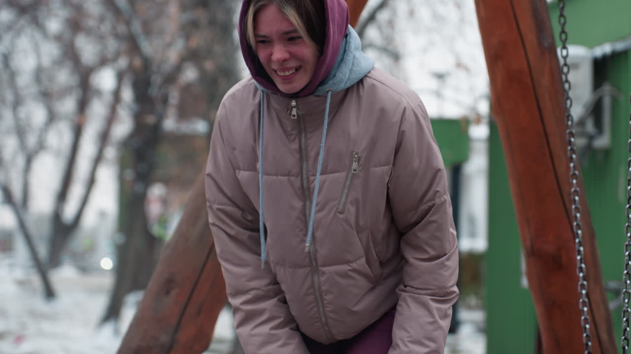 dama en traje de invierno sintiendo dolor en la rodilla mientras se inclina con árboles y fondo nevado, mostrando expresión de incomodidad, usando capucha y chaqueta hinchada, escena de invierno
