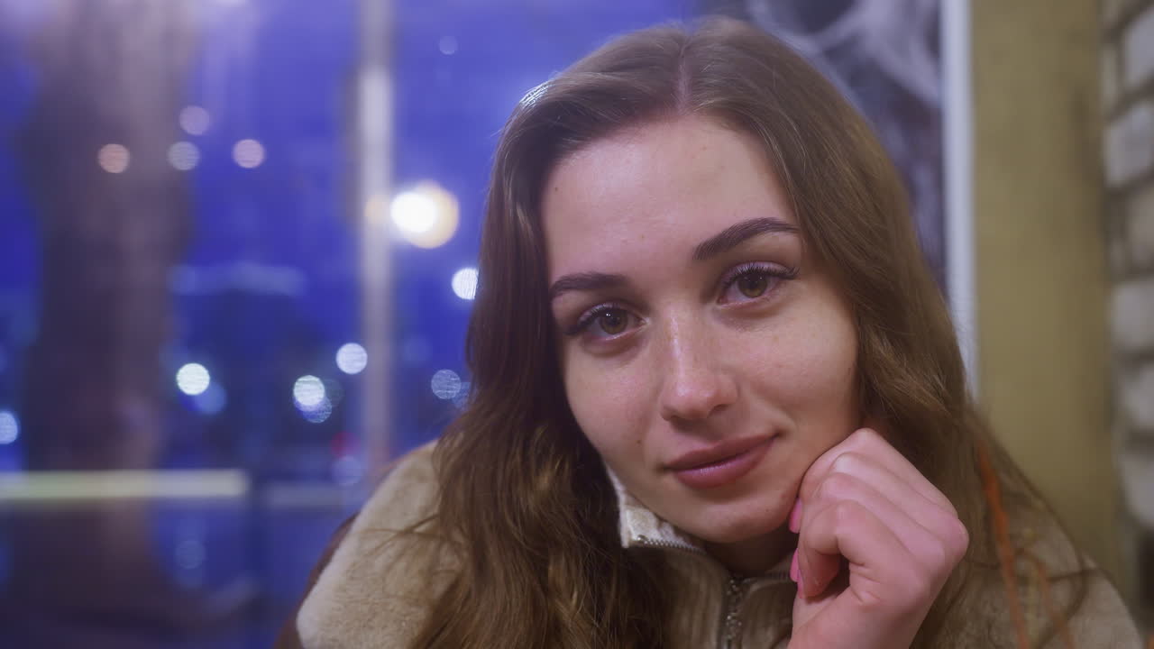 Close-up shot of young woman in brown shearling jacket sitting in cozy cafe, resting forward on table with relaxed and joyful expression. Moving cars visible through window in background