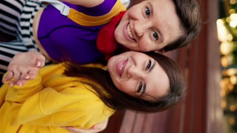 Vertical-video:-Happy-lesbian-couple-and-a-brunette-girl-in-a-yellow-sweater-and-a-girl-with-a-short-haircut-in-a-purple-top-and-red-headphones-take-a-selfie-and-rejoice-on-a-bench-in-a-skatepark-in-summer
