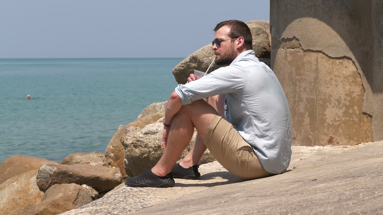 Hombre sentado en la playa sobre las rocas con una bebida, relajándose.