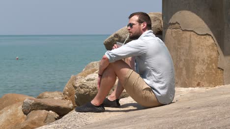 Mann-Sitzt-Am-Strand-Auf-Felsen-Mit-Einem-Entspannenden-Getränk