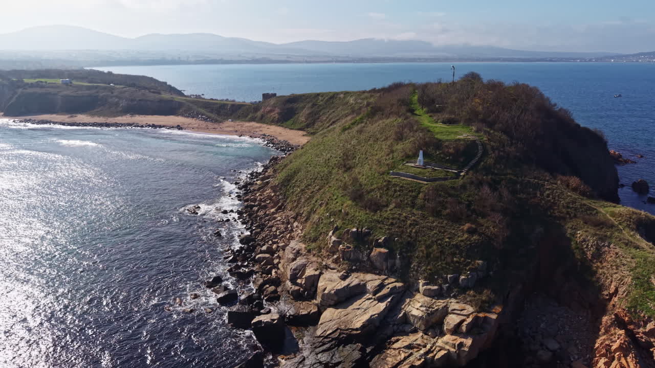 Naturskønt kystlandskab med klippefyldte kyster og blide bølger under en klar blå himmel nær en fredfyldt strand tidligt på eftermiddagen.