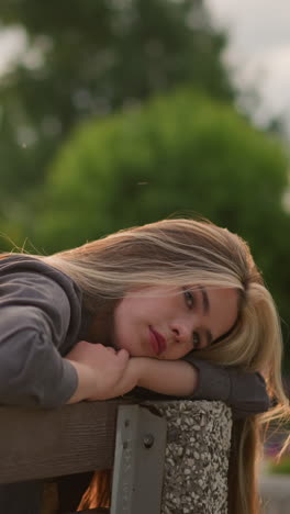 dama sentada en un banco en el borde, descansando la cabeza y los brazos con una expresión tranquila, mientras la luz del sol crea luz reflectante a su alrededor, el fondo presenta árboles con una vista borrosa de personas caminando