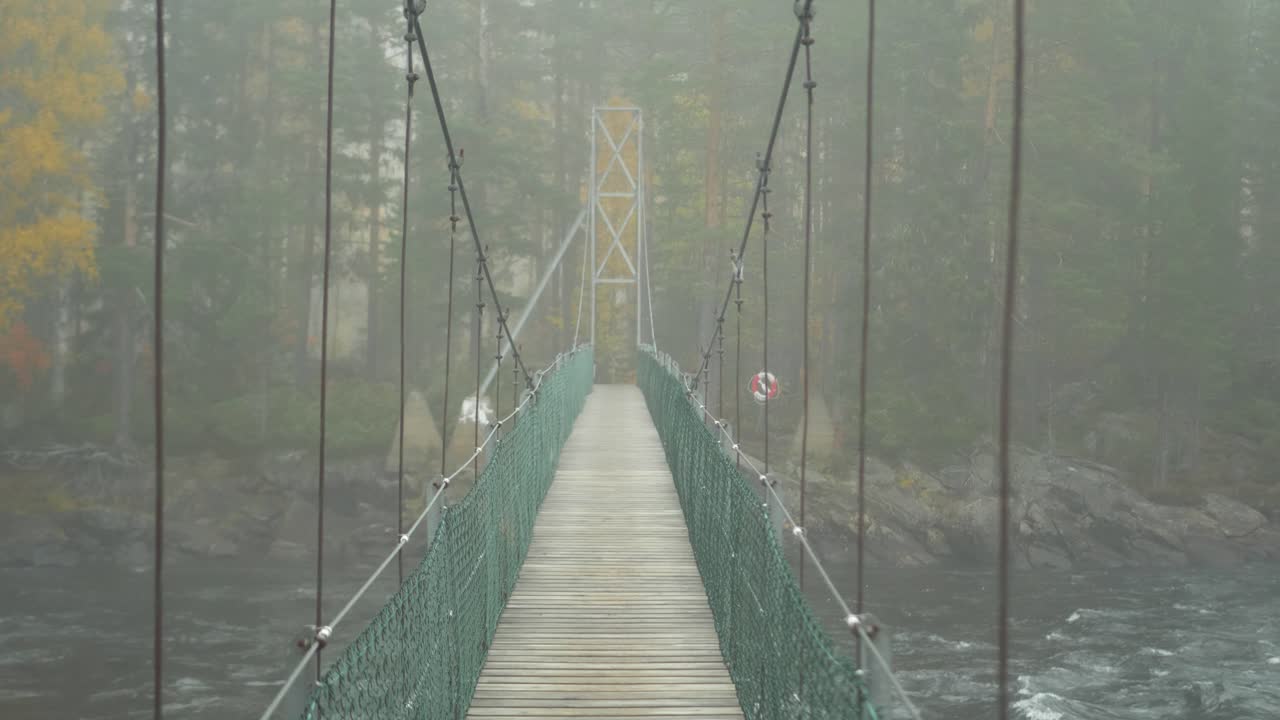 POV crossing Suspended Wooden Bridge over violent turbulent river on a misty forest