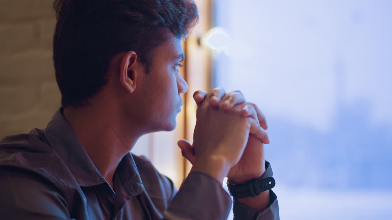 Young man sits indoors near window with hands clasped before face, staring out into bright blue background, conveying deep thought, quiet emotion, personal reflection, and peaceful stillness in natural light
