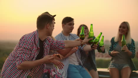 a man raises a toast to a friend's birthday in friends company on the roof. they clink beer and eat pizza.