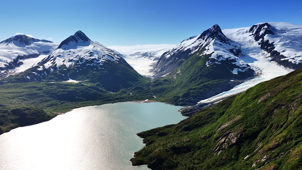Stunning mountains covered with snow reflecting sunlight. Lake at the mountain foot sparkles in the sun. Aerial view. Alaska, USA