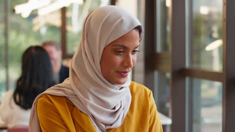 Mature-Businesswoman-Wearing-Headscarf-Working-At-Desk-In-Office-Pausing-To-Look-Out-Of-Window