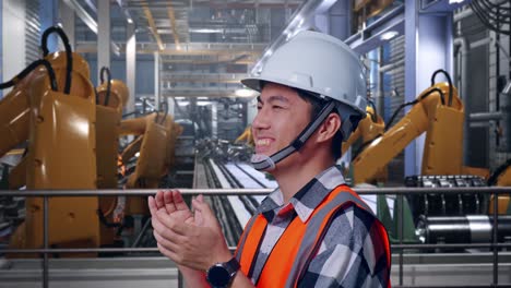 close up side view of asian male engineer with safety helmet smiling and clapping his hands while standing with industrial robotic arms working
