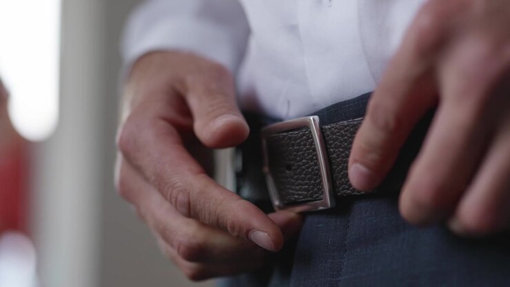 Close-up of groom fastening leather belt while getting ready for wedding ceremony. Elegant preparation detail highlighting formal attire and anticipation before marriage.