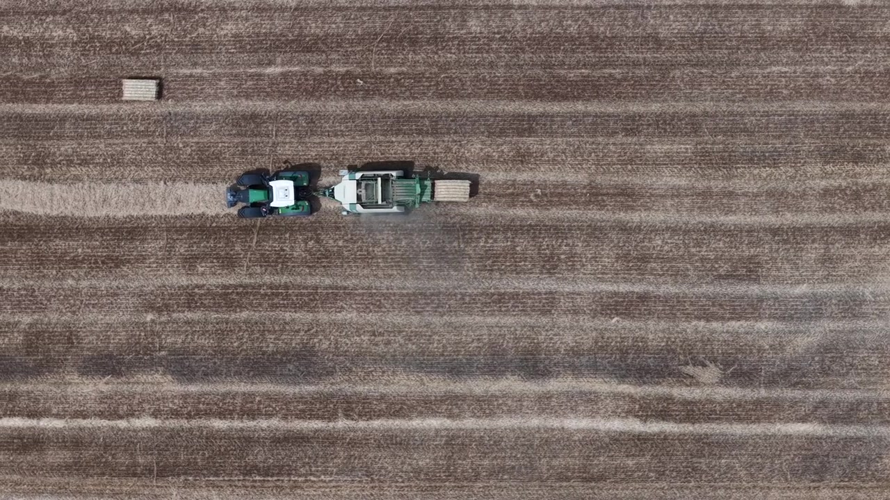 Un trattore e una pressa per balle producono efficientemente balle di fieno rettangolari in un vasto campo mietuto, una vista aerea che illustra le moderne pratiche agricole.
