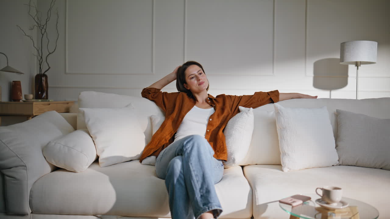 Happy woman lounging sofa modern living room. Relaxed brunette sit down couch