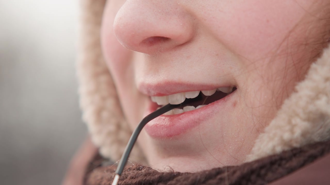 Primo piano frontale ravvicinato della parte inferiore del viso che indossa un cappuccio foderato di pelliccia e una sciarpa di maglia testurizzata, che tiene l'asta degli occhiali tra i denti con le labbra leggermente dischiuse, un sorriso sottile e un'atmosfera invernale rilassata all'aperto.