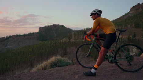 Atop-mountain,-sportsman-in-yellow-t-shirt,-helmet,-and-gear-rests-on-bike,-admiring-mountains-and-setting-sun-after-workout