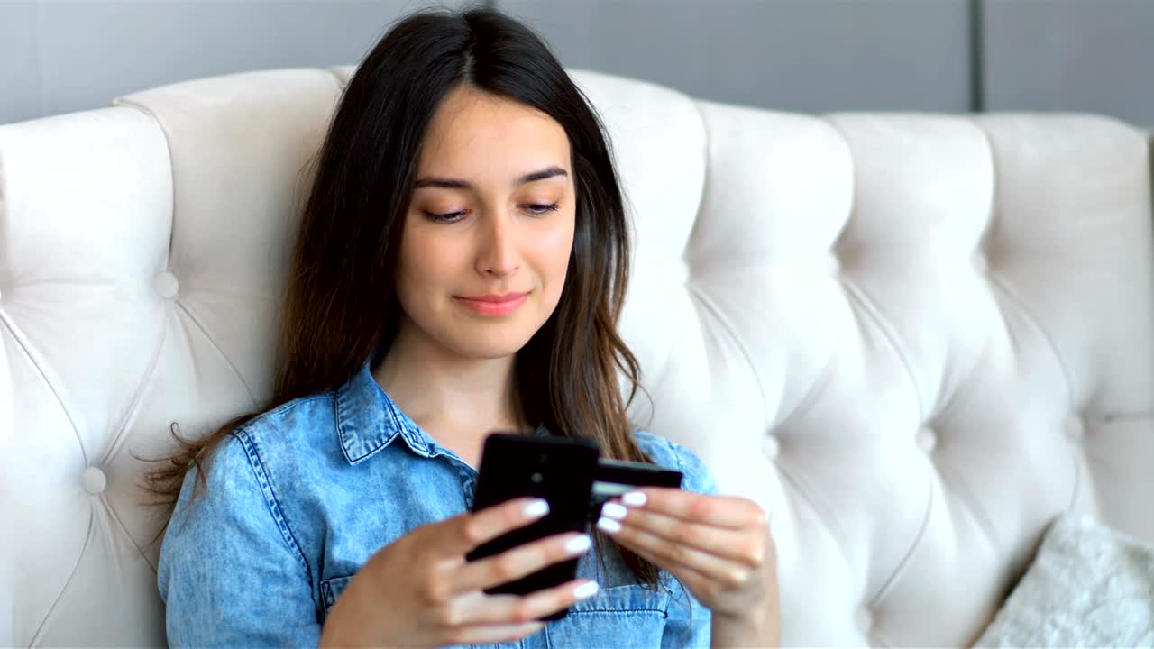joven bonita comprando en línea con tarjeta de crédito y teléfono inteligente sentada en un sofá en la sala de estar en casa