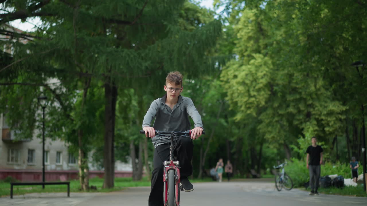 공원 지역을 통해 꾸준히 자전거를 타고 있는 어린 소년, 안경과 회색 을 입고, 배경에는 나무의 울창한 풍경, 도시 건물, 그리고 흐릿한 사람들이 고 있습니다.