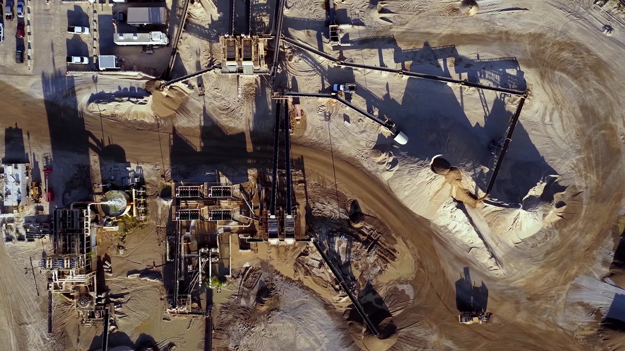 Drone video captures a straight-down, sliding aerial of an active gravel and sand pit at sunrise. Long shadows emphasize machinery, conveyors, and earth textures in crisp golden light.