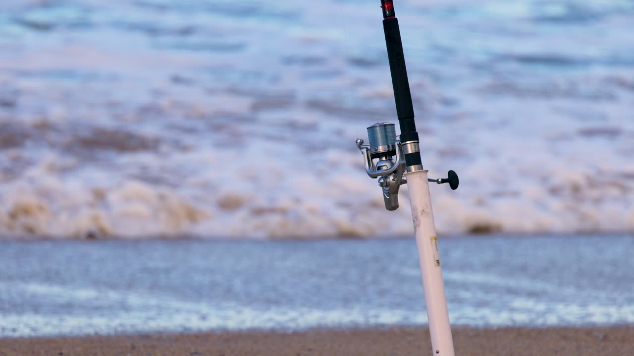 A solitary fishing rod stands on a sandy beach with waves gently rolling in under soft, natural lighting